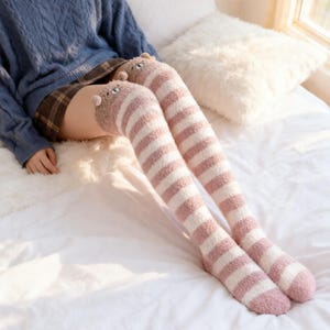 May include: Over-the-knee socks with a cat face design at the top. The socks are pink and white striped with a fuzzy texture. The socks are knee-high.