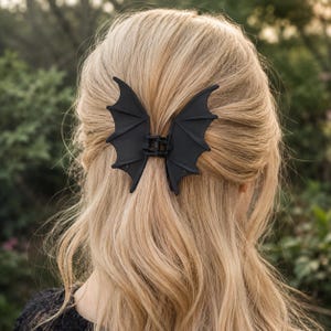 May include: A matte black hair claw clip shaped like a pair of bat wings. The accessory features a hinged mechanism in the center, allowing the two wing-shaped sides to open and grip hair securely. The design is symmetrical with pointed tips.