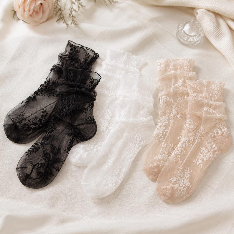 K&ouml;nnte beinhalten: Drei Paar transparente Kn&ouml;chelsocken mit zarter Blumenstickerei und ger&uuml;schten B&uuml;ndchen. Das Set enth&auml;lt ein schwarzes, ein wei&szlig;es und ein beiges Paar, die alle aus einem leichten, halbtransparenten Netzstoff gefertigt sind.