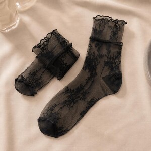 K&ouml;nnte beinhalten: Ein Paar transparente schwarze Kn&ouml;chelsocken mit einem zarten floralen Spitzenmuster auf dem gesamten Stoff. Die Socken haben einen ger&uuml;schten, gewellten Rand am oberen B&uuml;ndchen sowie einen verst&auml;rkten, einfarbig schwarzen Zehen- und Fersenbereich.
