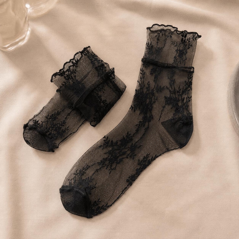 K&ouml;nnte beinhalten: Ein Paar transparente schwarze Kn&ouml;chelsocken mit einem zarten floralen Spitzenmuster auf dem gesamten Stoff. Die Socken haben einen ger&uuml;schten, gewellten Rand am oberen B&uuml;ndchen sowie einen verst&auml;rkten, einfarbig schwarzen Zehen- und Fersenbereich.