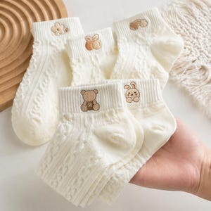 May include: Cream-coloured ankle socks with a cable knit texture. Each sock features a different embroidered design: a flower, a bunny, a cloud, a bear, and another bunny. The socks are held in a hand.