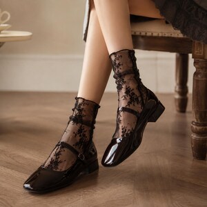 K&ouml;nnte beinhalten: Ein Paar schwarze Mary-Jane-Schuhe aus Lackleder mit niedrigem Blockabsatz und einem Riemen &uuml;ber dem Spann, getragen mit transparenten schwarzen S&ouml;ckchen mit filigranem Blumen-Spitzenmuster, die bis zum unteren Wadenbereich reichen.