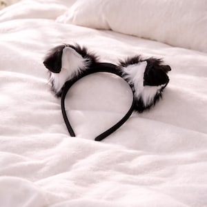 Pode incluir: Uma tiara preta com orelhas de gato fofas, pretas e brancas. As orelhas são feitas de pele sintética macia e têm formato pontudo. A tiara está sobre uma superfície branca texturizada.