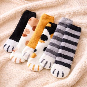 Peut inclure: Six paires de chaussettes douillettes et duveteuses avec des motifs de pattes de chat. Les chaussettes présentent diverses couleurs, dont le noir, le blanc, l'orange et le gris, avec des empreintes de pattes sur les orteils. Les chaussettes sont disposées sur une couverture crème.
