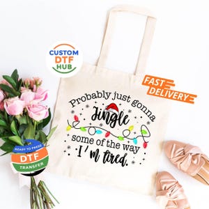 May include: A cream-colored tote bag with the text "Probably just gonna Jingle some of the way, I'm tired." The design includes a Santa hat, Christmas lights, and snowflakes. The bag is next to a bouquet of pink roses.