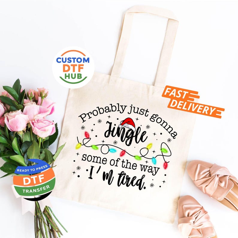 May include: A cream-colored tote bag with the text "Probably just gonna Jingle some of the way, I'm tired." The design includes a Santa hat, Christmas lights, and snowflakes. The bag is next to a bouquet of pink roses.