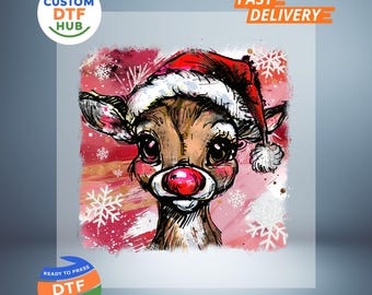 Op de afbeelding: Een grillige illustratie van een rendier met een kerstmuts, een felrode neus en roze wangen. De achtergrond heeft een roze en wit aquareleffect met sneeuwvlokaccenten. De afbeelding bevat de tekst "Custom DTF Hub" en "Ready to Press DTF Transfer".