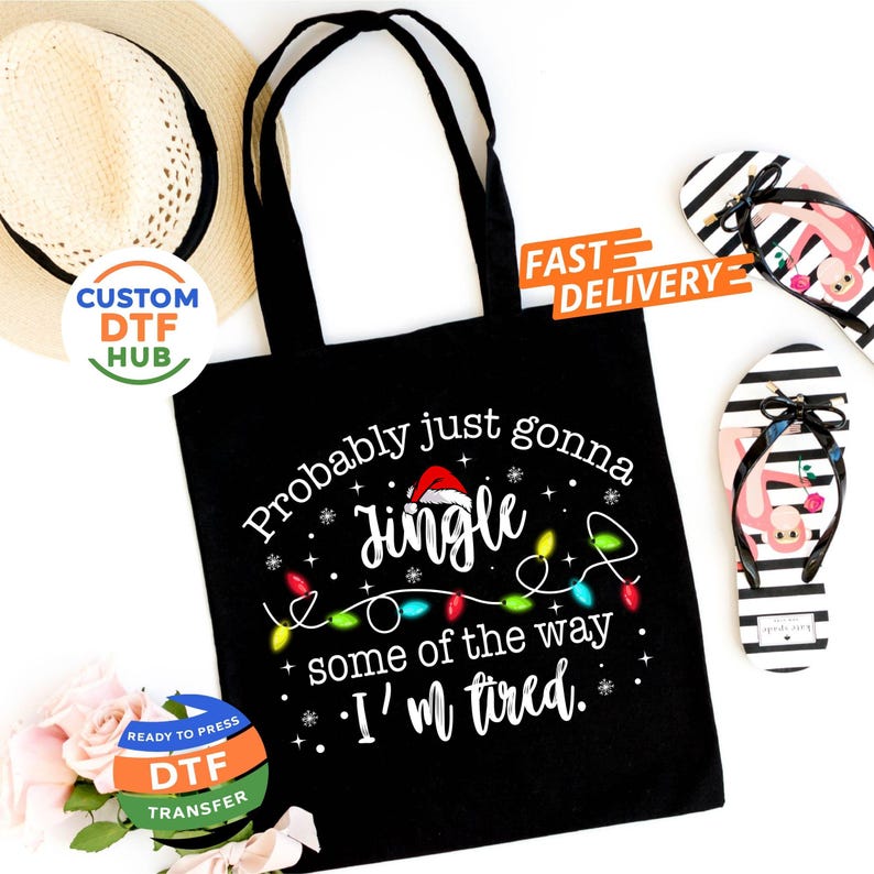May include: Black tote bag with the text "Probably just gonna Jingle some of the way, I'm tired." The design includes a Santa hat, Christmas lights, and snowflakes. Also shown are a straw hat and a pair of black and white striped flip-flops.