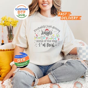 May include: Cream-colored t-shirt with the text "Probably just gonna jingle some of the way, I'm tired." The design includes a Santa hat, string lights, and stars. The shirt is part of a Christmas-themed collection.