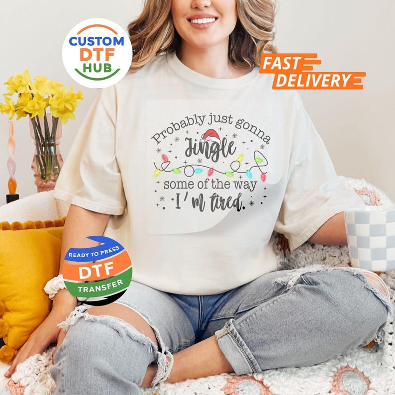 May include: Cream-colored t-shirt with the text "Probably just gonna jingle some of the way, I'm tired." The design includes a Santa hat, string lights, and stars. The shirt is part of a Christmas-themed collection.