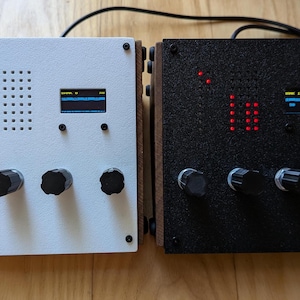 Puede incluir: Dos dispositivos electrónicos con marcos de madera. Uno es blanco con perillas negras y una pequeña pantalla, el otro es negro con perillas similares y una pantalla. Ambos tienen luces y están conectados por un cable negro.