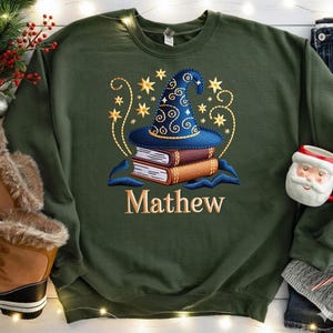 Może przedstawiać: Ciemnozielona bluza z nadrukiem niebieskiego kapelusza czarodzieja na dwóch książkach, otoczona złotymi gwiazdami i wirami. Imię "Mathew" jest wyszyte złotem. Świąteczny motyw.