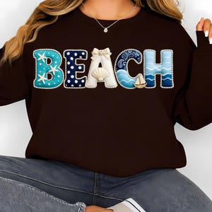 May include: A brown sweatshirt with the word "BEACH" in a decorative, beach-themed design. The letters are in various colors and patterns, including blue with white polka dots, a seashell, and a sailboat.
