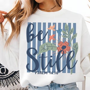 May include: White sweatshirt with a blue and white striped background design. The words "Be Still" are written in a script font, with floral and bird illustrations. The text "Psalm 46:10" is also included.