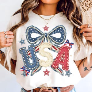 Op de afbeelding: Crèmekleurig T-shirt met een patriottisch USA-ontwerp. Het ontwerp heeft een denim-achtige strik met witte sterren en parelaccenten, samen met de letters "USA" in rood, wit en blauw, ook versierd met parels. Sterren en strikken maken het ontwerp compleet.