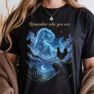 May include: A black t-shirt featuring a graphic print of a glowing blue spectral Tyrannosaurus Rex looming over a silhouette of a rooster standing on a cliff edge, with the text "Remember who you are." printed in gold above the dinosaur illustration.