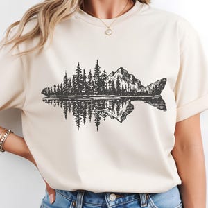 May include: A cream-colored short-sleeve t-shirt featuring a black graphic print of a fish silhouette filled with a detailed landscape of pine trees and a mountain peak, including a mirrored reflection effect of the scenery below the fish.