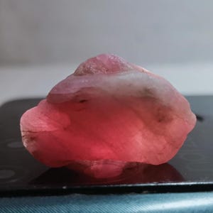 Peut inclure: Une pierre précieuse brute et non taillée d'une couleur rose vif. La pierre présente une surface rugueuse et texturée, ainsi qu'un aspect légèrement translucide. La forme est irrégulière, avec des bords arrondis et une forme naturelle. Il s'agit probablement d'un spécimen minéral.