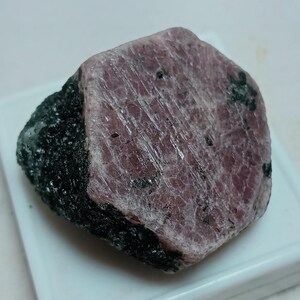 May include: A raw ruby gemstone with a deep red-pink hue, partially encased in a dark, rough matrix. The ruby's surface has visible striations and small black inclusions, showcasing its natural crystalline structure. This uncut gemstone is a unique specimen.