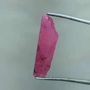 May include: A raw, uncut ruby gemstone, held by tweezers. The ruby is a vibrant pinkish-red color with a rough, textured surface. Small dark inclusions are visible within the stone. The gemstone is displayed against a neutral background.