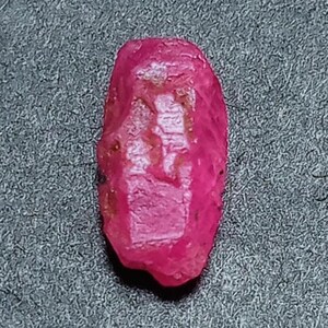 Puede incluir: Una gema de rubí natural, en bruto. El rubí es de un color rosa rojizo intenso y vibrante, con una forma ligeramente irregular. La superficie tiene un aspecto texturizado, con inclusiones e imperfecciones naturales visibles. La gema se encuentra sobre un fondo gris.