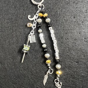 May include: A silver keychain featuring two beaded strands with black, grey, and gold beads. One strand spells out TO WHATEVER END with letter cubes. Charms include a crescent moon, a book, a sword, a feather, and a silver deer figurine.