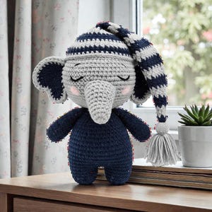 May include: A handmade crochet elephant doll wearing a navy blue and white striped nightcap and a navy blue onesie. The elephant has gray skin, large ears, and a long trunk. The doll has closed eyes and pink cheeks, suggesting it is sleeping.