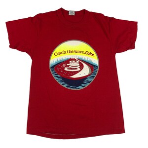 May include: A red t-shirt featuring a circular graphic with the text "Catch the wave. Coke." The graphic depicts a stylized ocean scene with the word "Coke" in white script. The shirt is a classic crew neck style.