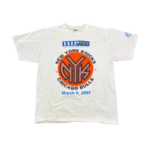 May include: A white short-sleeve t-shirt featuring a graphic with the text HIP 50 ANNIVERSARY 1947-1997, NEW YORK KNICKS, CHICAGO BULLS, and March 9, 1997. The central design includes a large orange circle with the letters NYK in black and red.