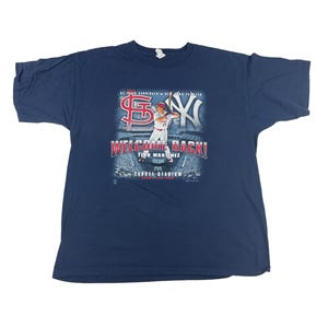 以下が含まれることがあります： 赤いユニフォームを着てバットを持った野球選手のグラフィックがプリントされたネイビーブルーのTシャツ。グラフィックには「Welcome Back!」と「Tino Martinez」の文字が書かれています。