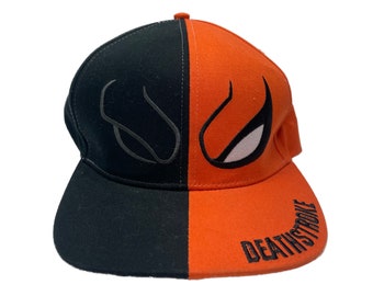 Könnte beinhalten: Eine zweifarbige Baseballkappe mit einer schwarzen linken Seite und einer orangefarbenen rechten Seite. Die Vorderseite zeigt ein gesticktes Augendesign mit einer dunklen Umrandung auf der schwarzen Seite und einem weiß gefüllten Auge auf der orangefarbenen Seite. Das Wort DEATHSTROKE ist schwarz auf den orangefarbenen Schirm gestickt.