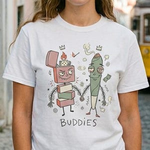 May include: White t-shirt featuring a cartoon illustration of a lighter and a cannabis joint holding hands, with the word "Buddies" below. The design includes flames, smoke, and crowns. The model is wearing blue jeans.