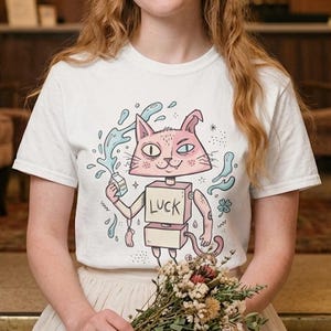 Pu&ograve; includere: T-shirt bianca con un disegno di gatto dei cartoni animati. Il gatto rosa ha dettagli blu e la parola "LUCK" sul corpo. Tiene in mano un oggetto con schizzi d'acqua intorno. La persona che indossa la maglietta tiene in mano un mazzo di fiori secchi.