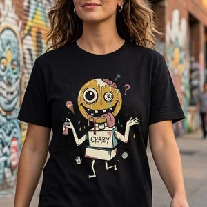 May include: Black t-shirt with a cartoon character design. The character has a yellow head, a brain, and a long tongue. The word "CRAZY" is printed on the character's body. The background shows graffiti art and a brick wall.