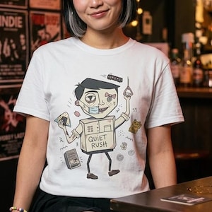 Op de afbeelding: Wit T-shirt met een cartoonfiguur met een doosvormige torso en de tekst "QUIET RUSH". Het personage houdt een metroband en een sandwich vast. Concertposters zijn op de achtergrond.