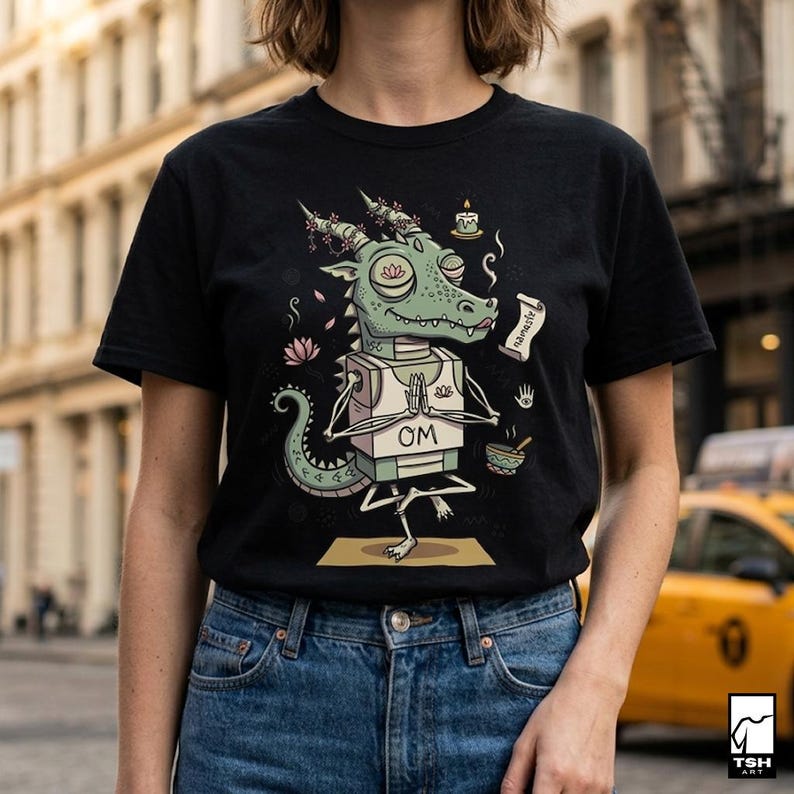 Op de afbeelding: Zwart t-shirt met een cartoon draak in een yoga pose. De groene draak heeft een witte doos met "OM". Het ontwerp bevat een kaars, lotusbloemen en het woord "Namaste".
