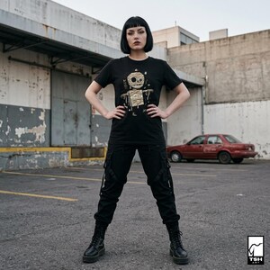 Puede incluir: Camiseta negra con un dise&ntilde;o de momia y la palabra "Cursed" en dorado. La persona lleva pantalones cargo negros y botas negras. Un coche rojo y un edificio de hormig&oacute;n se ven al fondo.