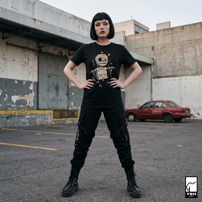 Puede incluir: Camiseta negra con un dise&ntilde;o de momia y la palabra "Cursed" en dorado. La persona lleva pantalones cargo negros y botas negras. Un coche rojo y un edificio de hormig&oacute;n se ven al fondo.