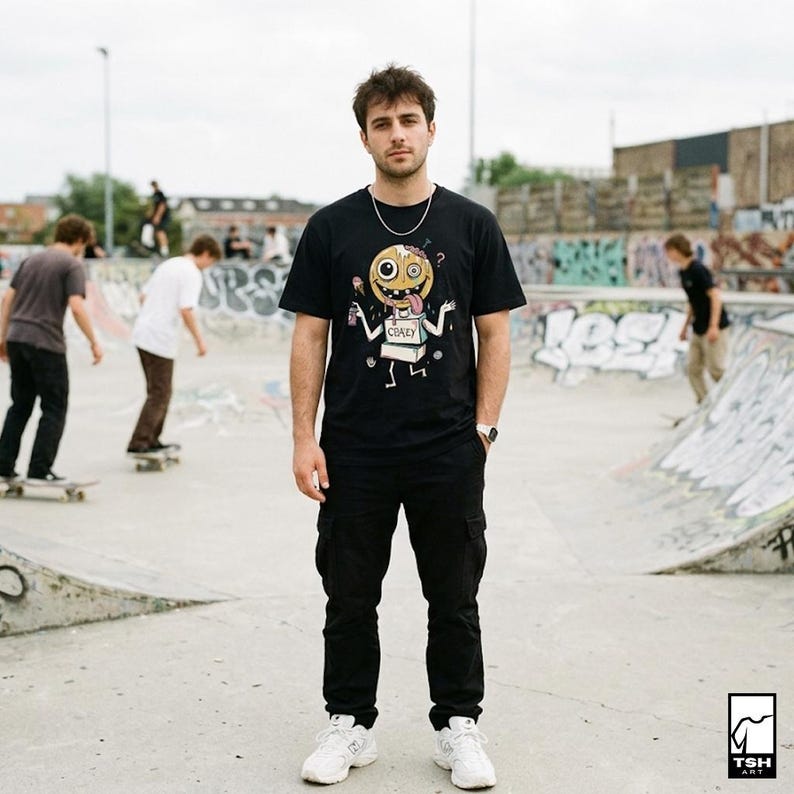 May include: A person wearing a black t-shirt with a cartoon graphic and the word "CRAZY". The person is standing in a skate park, wearing black trousers and white trainers. Other people are skateboarding in the background.