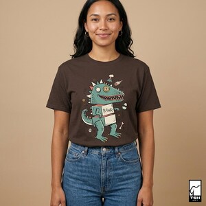May include: A brown t-shirt featuring a cartoon dinosaur with the word "RAWR" on a box it is holding. The dinosaur is teal with a party hat and other space-themed elements. The shirt is paired with blue jeans.
