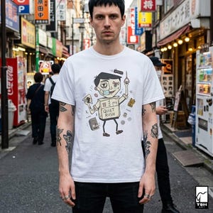 Op de afbeelding: Wit t-shirt met een cartoon illustratie van een persoon in een doos, met een gezichtsmasker en een kaartje en een schaar. De tekst "QUIET RUSH" staat op de doos. De persoon staat op straat.