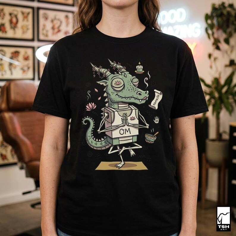 Op de afbeelding: Zwart T-shirt met een cartoon draak in een yoga pose. De draak is groen met een witte borst en het woord "OM" erop gedrukt. Het ontwerp bevat een lotusbloem, een kaars en een rol.