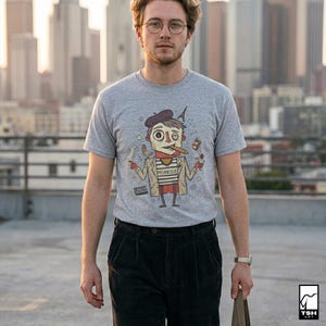 May include: A gray t-shirt featuring a cartoon illustration of a person with a beret, cigar, and the Eiffel Tower. The word "MELANCOLIE" is printed on the shirt. The person is wearing black pants and a watch. The background shows a cityscape.