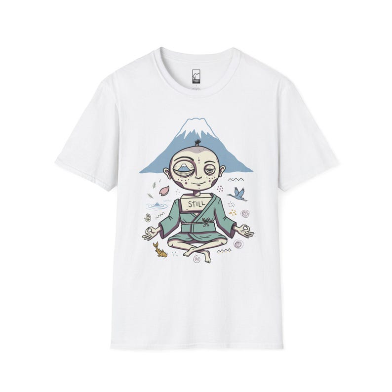 May include: A white short-sleeve t-shirt featuring a graphic of a figure in a teal robe sitting in a meditative pose. The figure has a box on their chest labeled STILL and a mountain in the background with small icons of a fish, bird, and petals.