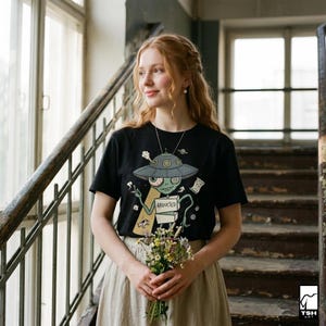 Może przedstawiać: Czarny t-shirt z kr&oacute;tkim rękawem z kresk&oacute;wkowym motywem kosmity. Kosmita, w szarej czapce statku kosmicznego, trzyma znak z napisem "ABDUCTED".