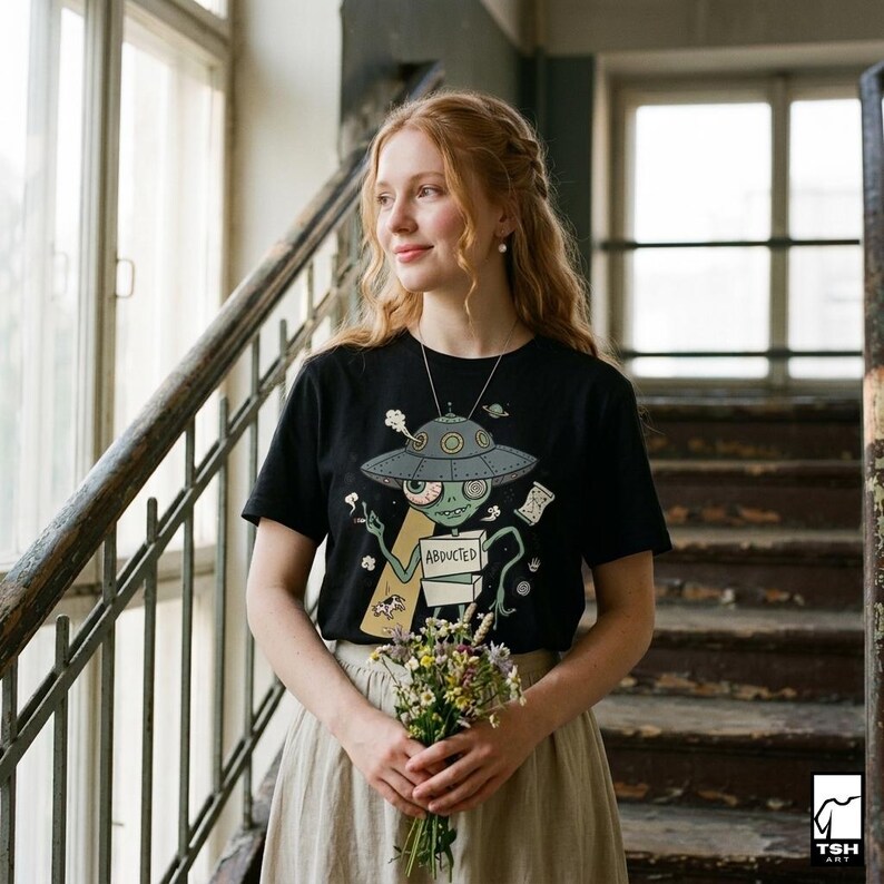 Może przedstawiać: Czarny t-shirt z kr&oacute;tkim rękawem z kresk&oacute;wkowym motywem kosmity. Kosmita, w szarej czapce statku kosmicznego, trzyma znak z napisem "ABDUCTED".