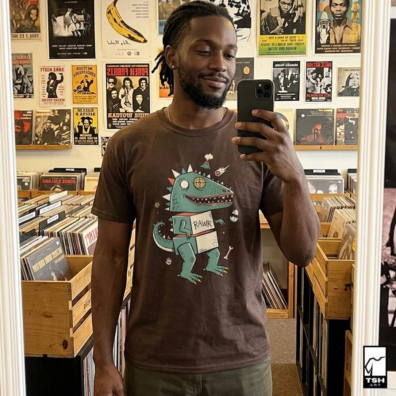 May include: A brown t-shirt featuring a cartoon dinosaur with a party hat and the word "RAWR" on a box. The dinosaur is teal and green with white accents. The person in the image is smiling and holding a phone.