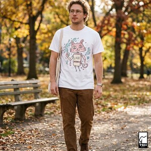 Pu&ograve; includere: T-shirt bianca con un disegno di un gatto dei cartoni animati che tiene in mano un cartello con la scritta "LUCK". Il gatto &egrave; rosa e ha uno spray blu intorno. La persona indossa pantaloni marroni e un orologio. Lo sfondo &egrave; un parco con alberi.