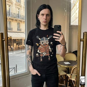 May include: Black t-shirt featuring a cartoon Viking playing a guitar, with the word "RAID" on the shirt. The image also shows a person wearing the shirt, black jeans, and holding a phone. The background includes a mirror and a window.